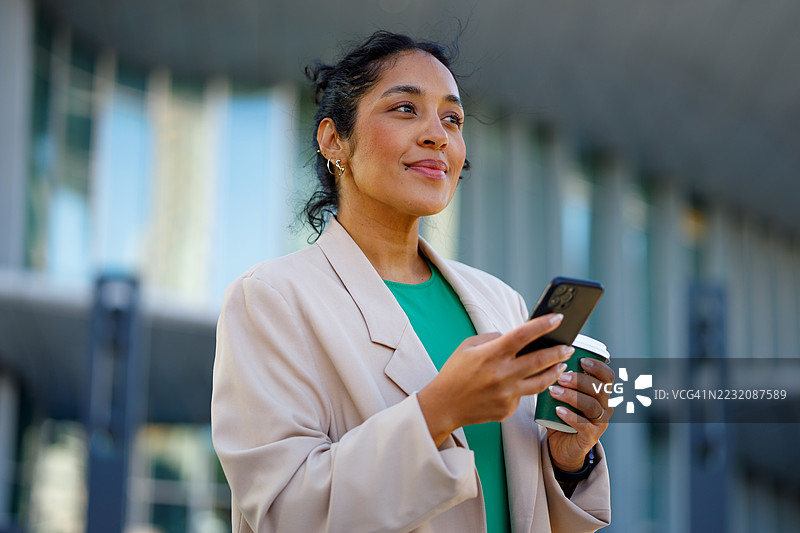 职业女性在现代玻璃办公大楼附近的城市环境中喝咖啡，使用智能手机工作。图片素材