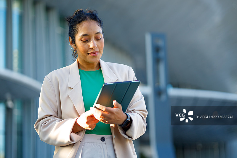 专业女商人在现代办公大楼旁站立，手握数字平板，自信地微笑着，身处城市环境中。图片素材