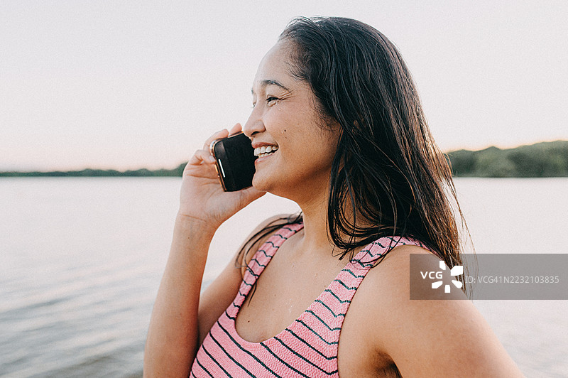 一位女性站在宁静的湖边，愉快地进行电话交谈。图片素材