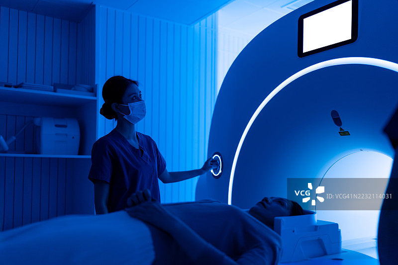 年轻的亚洲女性护士在MRI扫描室操作MRI扫描仪。女性医疗工作者在医疗扫描室内。蓝色调。图片素材