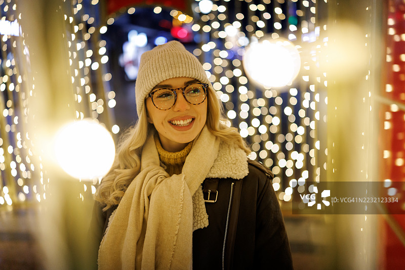快乐美丽的年轻女性在城市夜晚的圣诞灯光中愉快地微笑，享受节日气氛。图片素材