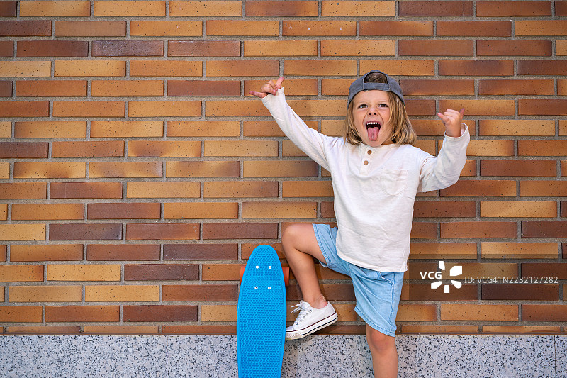 时尚的小孩拿着滑板，吐着舌头，做出摇滚手势，自信地站在充满活力的砖墙前，散发着顽皮的活力和个性。图片素材