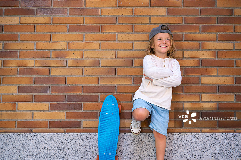 年轻、自信的滑板少年双臂交叉，倚靠在砖墙上，展示着一块蓝色滑板，微笑着望向一旁，散发出无忧无虑和酷炫的气息。图片素材