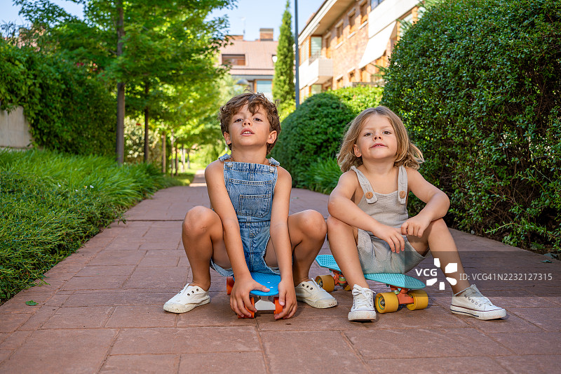 两个学龄前儿童蹲在五颜六色的塑料滑板上，在一个充满活力的公园里微笑着，直视镜头，享受阳光明媚的快乐友谊日。图片素材