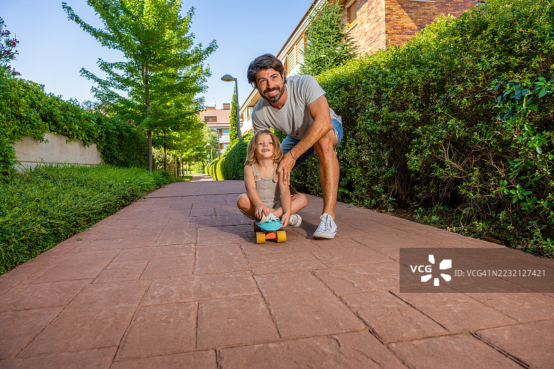 父亲微笑着教他的小儿子在住宅公园里滑滑板，营造出温馨的家庭亲密和户外乐趣的场景。图片素材