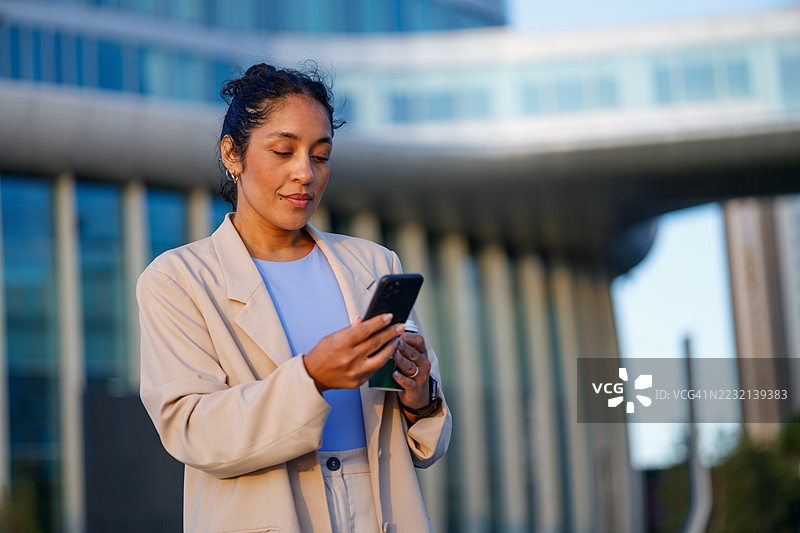 一位女商人在办公楼外使用智能手机，同时手里拿着咖啡杯，体现了现代都市生活方式。图片素材