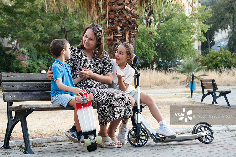 母亲和孩子们坐在公园的长椅上，身边有滑板和滑板车。图片素材