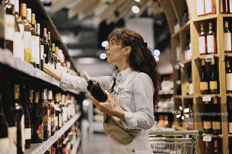 年轻女性在超市货架上挑选葡萄酒。她认真地检查一瓶酒，周围是摆满各种葡萄酒的货架。这个场景捕捉了现代杂货店中决策和日常生活的瞬间。图片素材