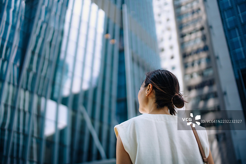 年轻的亚洲女商人在金融区低角度仰望，站在城市办公楼外墙前。看起来积极向上，充满动力。成功的女性领导力。职场女性。职业机会。决心追求成功。图片素材