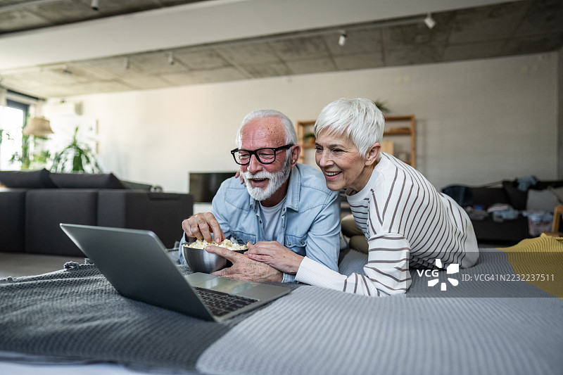 快乐的老年夫妇在家里看笔记本电脑上的电影，吃着爆米花图片素材