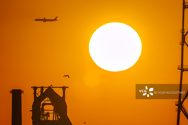 第一缕阳光与一架客机和鸟的剪影。它即将降落在附近的羽田机场。第一缕阳光从运河对岸的烟囱和电厂、钢铁厂上升。神奈川县横滨市，图片素材