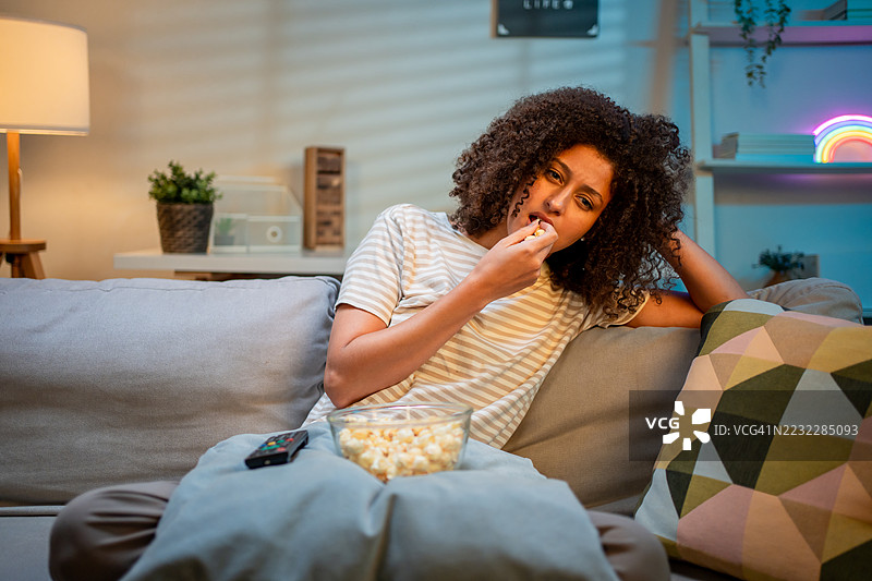 无聊的拉丁裔女性在家中夜晚看电视电影。迷人的女孩坐在沙发上，感到疲惫和无趣，观看电视系列节目。生活方式活动概念。图片素材