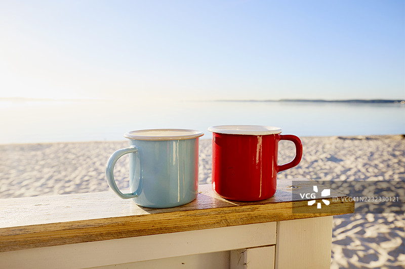 两个搪瓷咖啡杯放在海滩小屋的栏杆上，背景是日出时的海滩和大海。图片素材