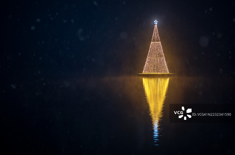 一棵完全照亮的漂浮圣诞树在夜晚的宁静湖面上闪耀着明亮的光芒，伴随着雪花飘落。图片素材