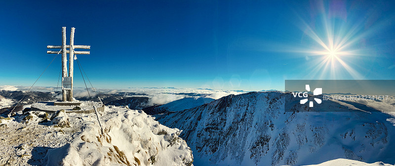 背光的全景冬季场景，山顶十字架和阳光照耀在被雪覆盖的山峰上，适合滑雪旅行。令人惊叹的阿尔卑斯山景，展现了高海拔冬季冒险的美丽与宁静。图片素材