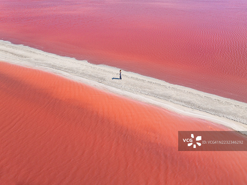 一张航拍照片，展示一位女性独自站在穿越粉色盐湖的小径上，法国卡马格。图片素材