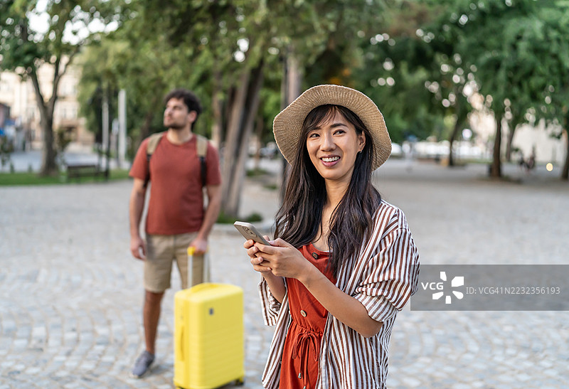 年轻游客情侣在城市中走动，拖着行李箱和背包，查看智能手机。图片素材