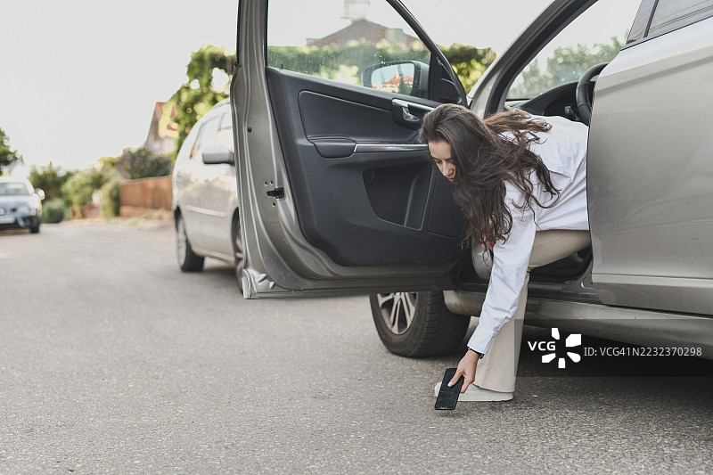 焦虑的女性从车内探身，仔细查看掉落在人行道上的碎屏手机图片素材