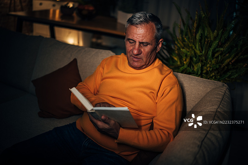 一位老人在安静的夜晚，舒适的客厅里阅读一本书。图片素材