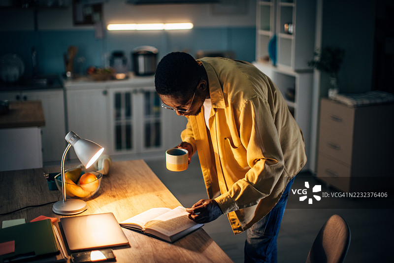年轻的黑人男子在温馨的夜晚氛围中，手捧咖啡阅读。图片素材