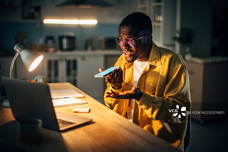 年轻的黑人男子在家中夜晚与人交谈，周围温暖的灯光照耀着他，专注于他的笔记本电脑。图片素材