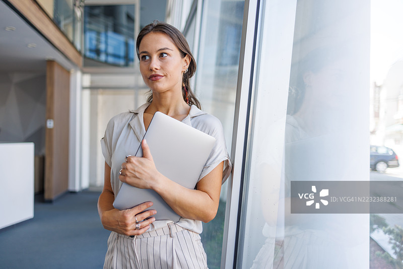 女商人在办公室走廊中手持笔记本电脑并侧目而视图片素材