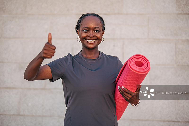 年轻的运动女性手持瑜伽垫，竖起大拇指。图片素材