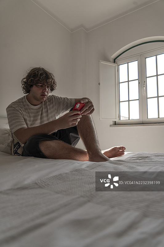 一个年轻男子坐在卧室的床上，在柔和的灯光下玩着手机，窗外的自然光洒了进来。图片素材