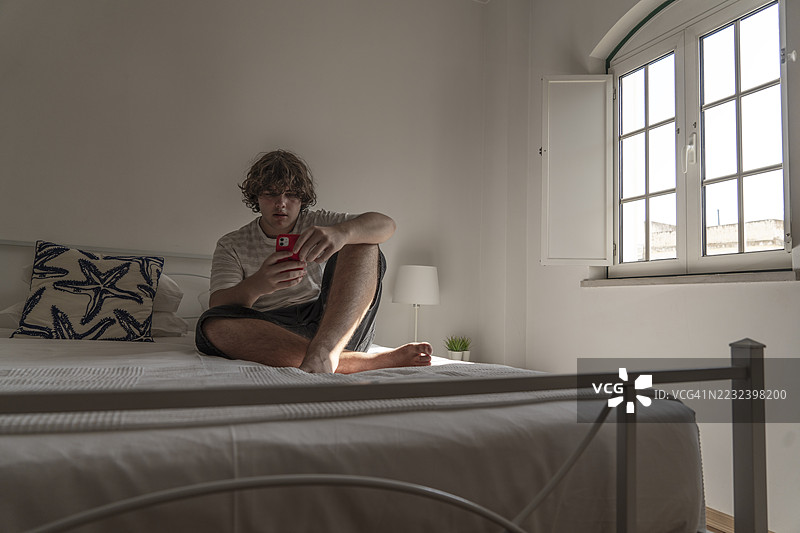 年轻男子在明亮的卧室里，午后阳光下使用智能手机放松图片素材