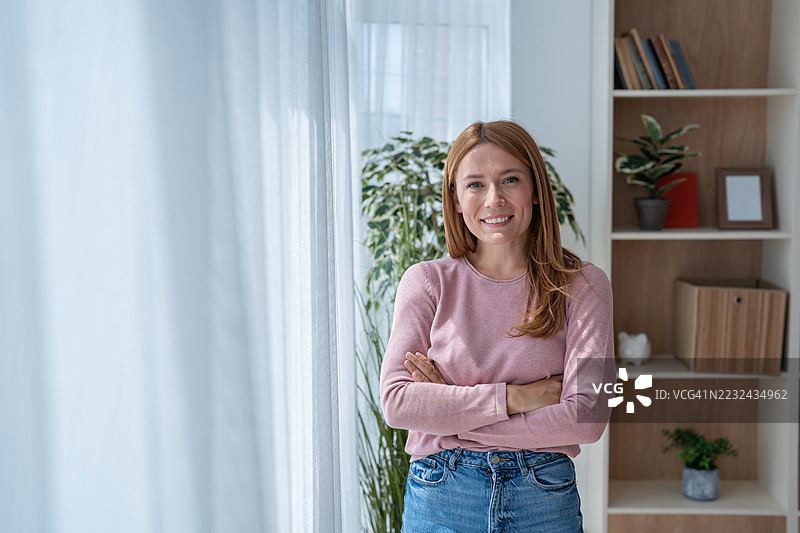自信的年轻女性微笑着，双臂交叉，站在家中的窗边。图片素材