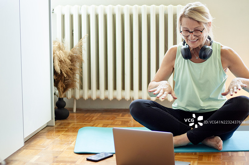 成熟的金发女性在公寓的瑜伽垫上使用笔记本电脑进行在线锻炼图片素材