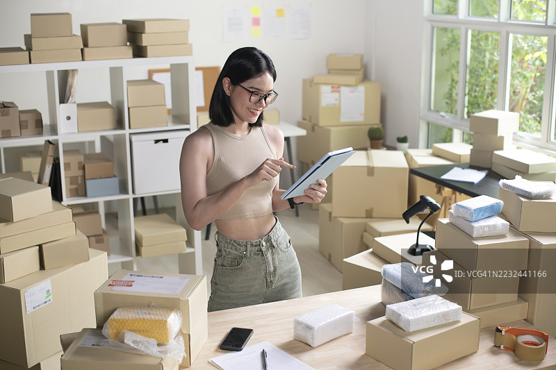 年轻女性在线卖家使用平板电脑准备包裹。代发货业务的店主在她的仓库工作。图片素材