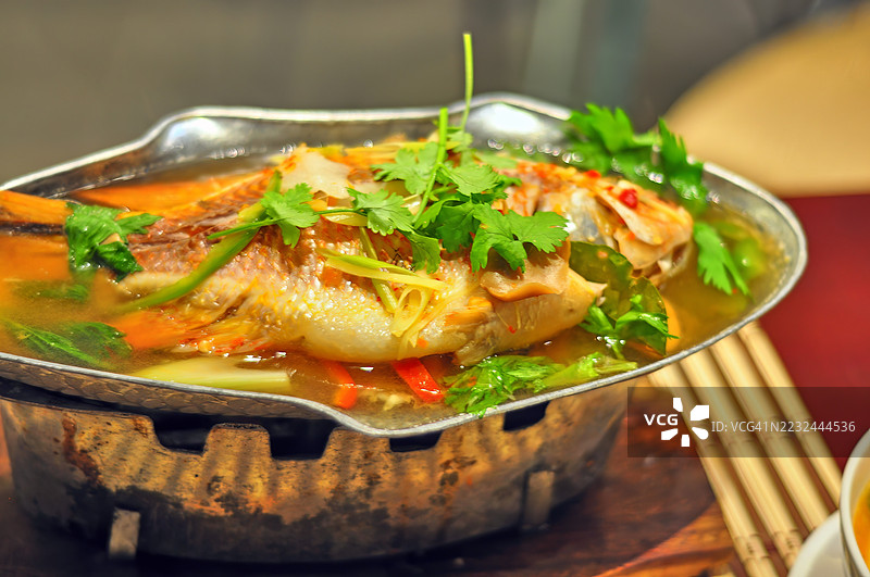 一只整条蒸鱼的特写俯视图，盛放在清汤中，放在一个不锈钢鱼形盘子上，置于保温器上。图片素材