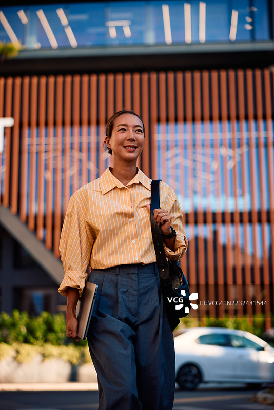 微笑的女商人在城市中走着，手里拿着笔记本电脑和包。图片素材