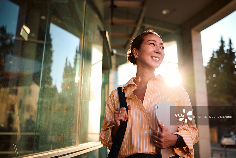 年轻的亚洲女商人在办公楼附近走着，手里拿着平板电脑，面带微笑。图片素材