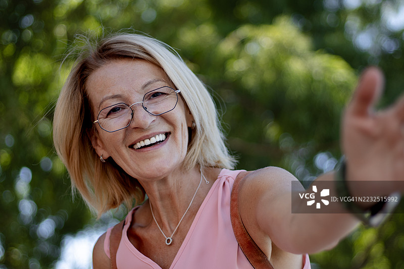 快乐微笑的成熟金发女性戴着眼镜，在大自然中享受美好时光的自拍照图片素材