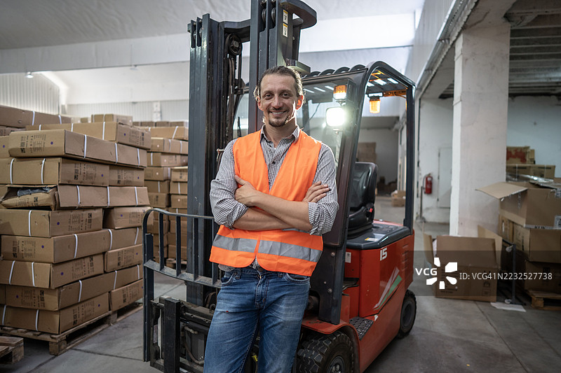仓库工人自信地站在叉车旁，周围是纸箱。图片素材