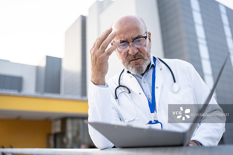 担忧的医生在现代医疗建筑外查看笔记本电脑数据图片素材