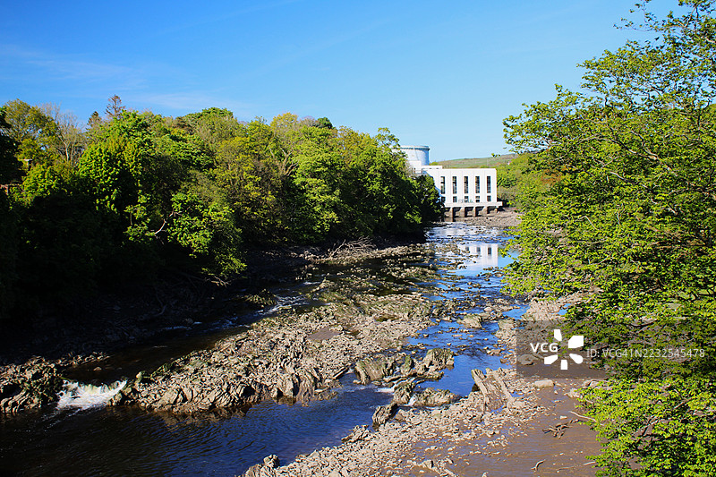 河流穿过郁郁葱葱的森林，背景中有一座建筑，克尔库德布赖特，英国图片素材