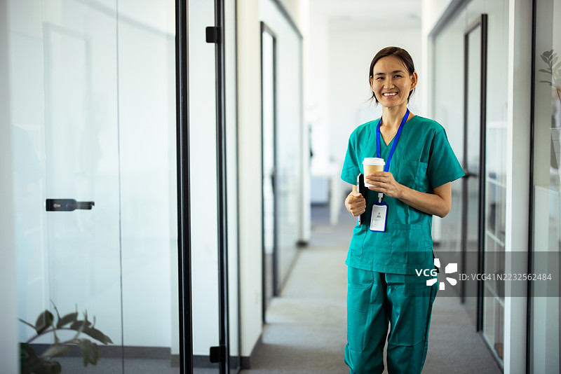 微笑的亚洲女性医生走在医院走廊里，手里拿着咖啡和平板电脑。图片素材