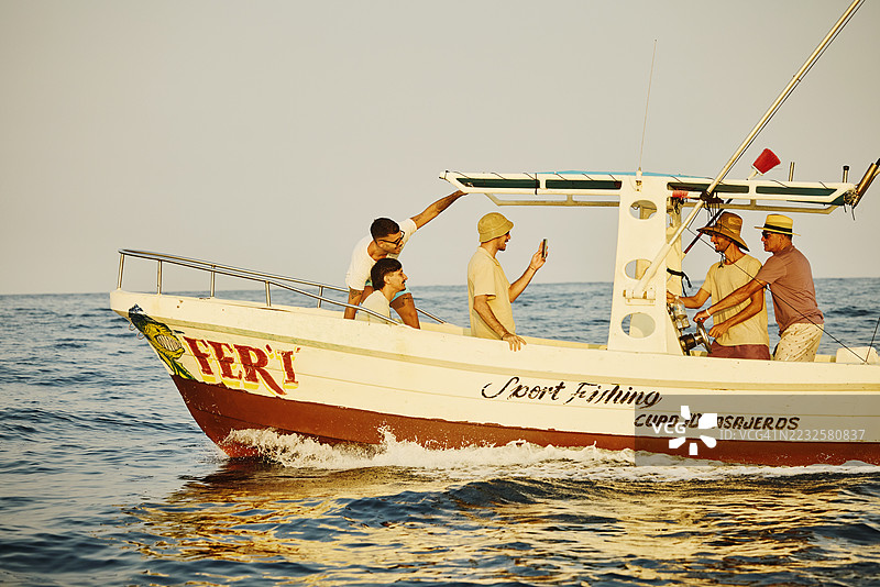 宽镜头拍摄儿子在运动钓鱼船上给爸爸和朋友拍照图片素材