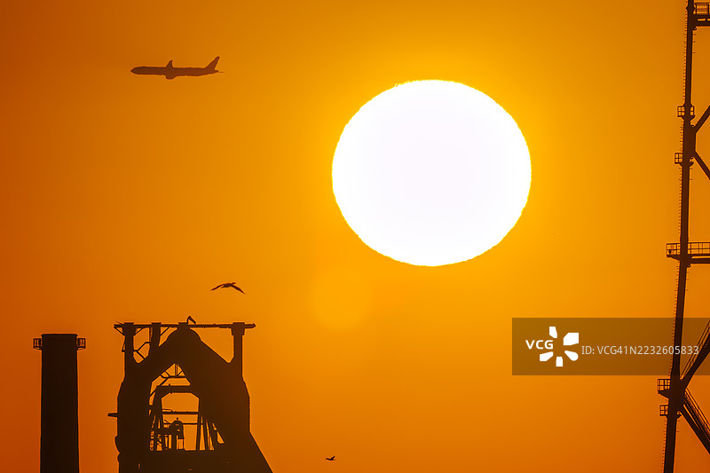 第一缕阳光与一架客机和鸟的剪影。它即将降落在附近的羽田机场。第一缕阳光从运河对岸的烟囱和电厂、钢铁厂上升。神奈川县横滨市，图片素材