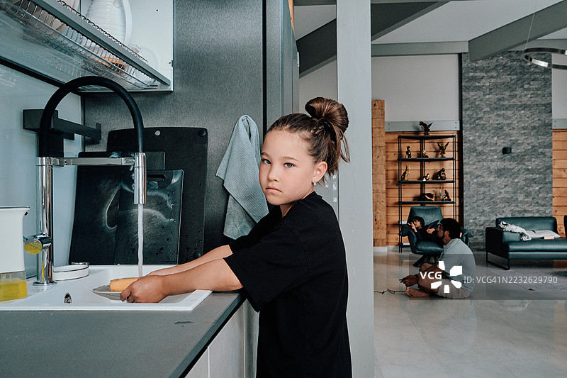 一个女孩在洗碗，父亲和儿子在背景中放松。图片素材