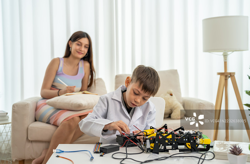 年轻男孩穿着实验室外套，正在组装机器人汽车，母亲在旁观看，STEM教育，儿童机器人工程，动手学习，创意活动，科学与技术图片素材