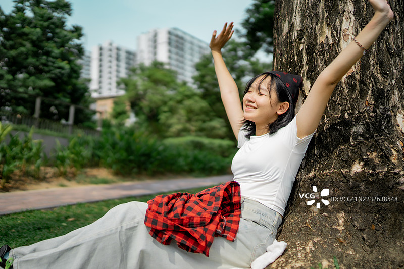 一位自信的年轻女孩，在绿意盎然的公园里靠着大树坐着，笑容灿烂，放松并享受自由，以快乐、俏皮和无忧无虑的户外活力拥抱这个广阔的世界，置身于大自然之中。图片素材
