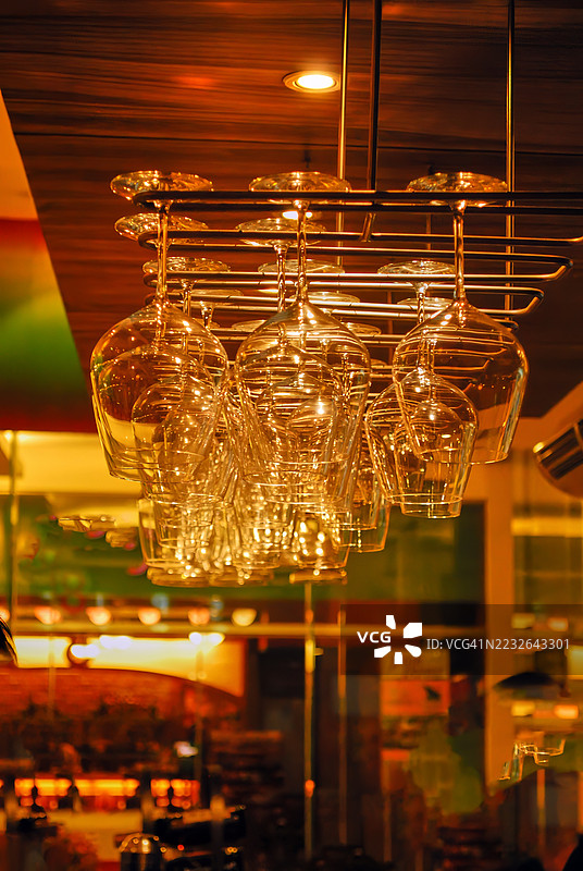 一排透明的酒杯倒挂在金属架上，置于温暖灯光的室内空间中。图片素材