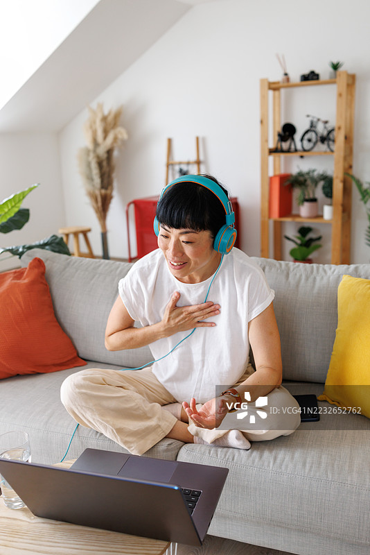 中年亚洲女性戴着耳机，在家沙发上通过笔记本电脑进行视频通话聊天图片素材