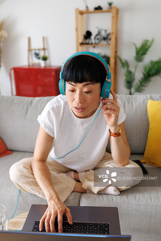 中年亚洲女性戴着耳机，在家沙发上通过笔记本电脑进行视频通话聊天图片素材