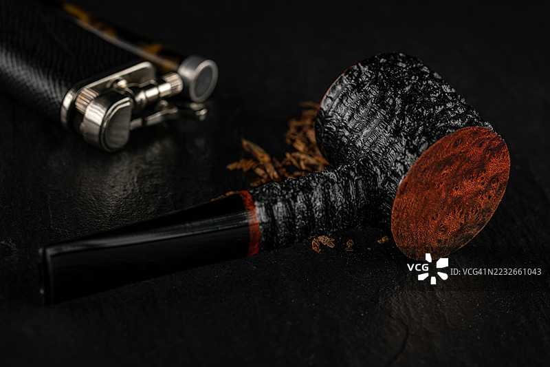 美丽的乡村黑色烟斗装满烟草，放置在石板上，背景有打火机和压烟器。图片素材