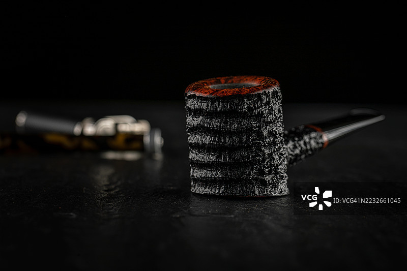 美丽的乡村黑色烟斗装满烟草，放置在石板上，背景中有打火机和压烟器。图片素材